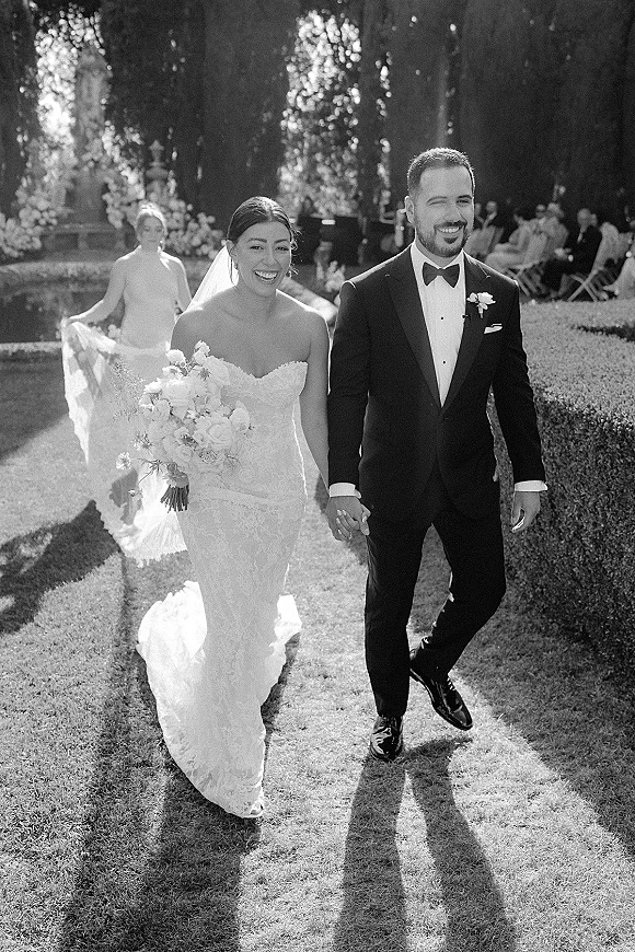 Wedding recessional with bride and groom walking hand in hand, bride in strapless lace gown and veil holding bouquet on sunlit garden lawn