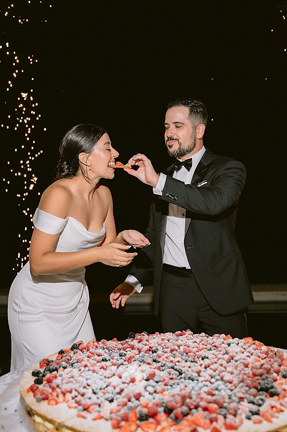 Wedding cake cutting as bride and groom share a bite beside a berry-topped cake with powdered sugar, sparklers glowing against the night sky