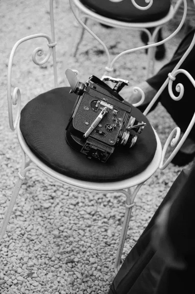 Film camera detail resting on a metal bistro chair with round cushion, vintage wedding photographer gear on gravel beside dark fabric