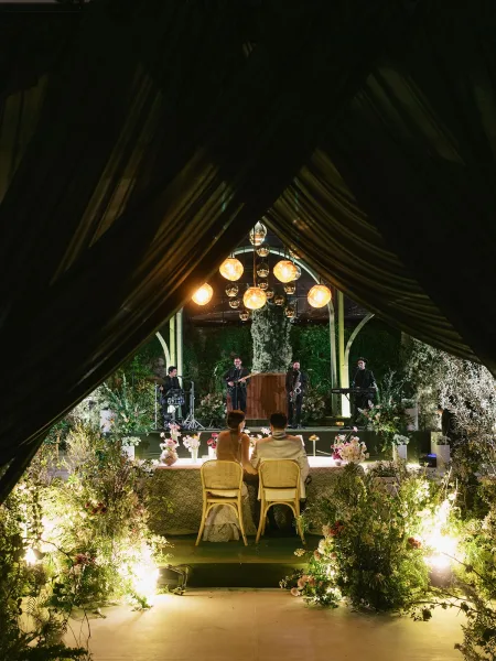 Wedding reception stage with live wedding band under draped ceiling fabric, hanging globe lights, and floral-lined platform in a garden at night