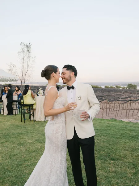 Couple portrait of bride and groom toast, laughing with champagne flute and rocks glass at vineyard cocktail hour in pastel sunset light