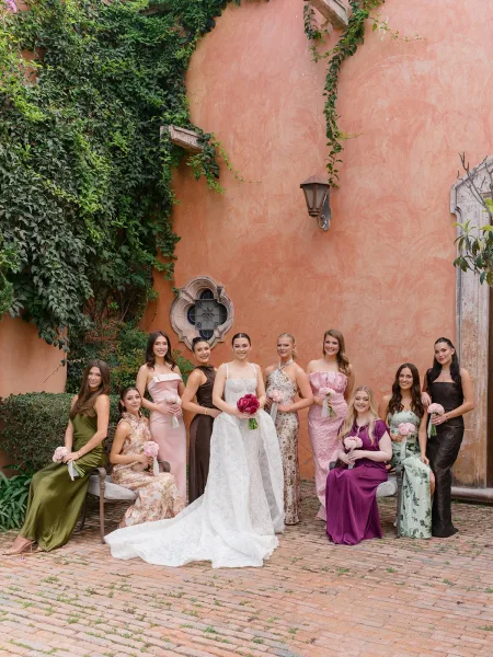 Bridal party portrait of the bride with bridesmaids in mismatched dresses holding bouquets by an ivy-covered stucco wall in a brick courtyard