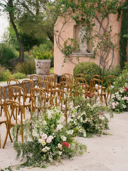 Ceremony setup with wood chairs lining an outdoor ceremony aisle, featuring aisle florals and greenery garland on a stone terrace courtyard