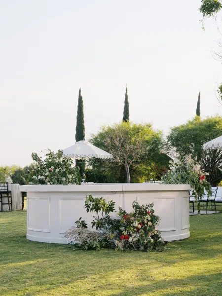 Outdoor wedding bar with a white paneled front and lush floral accents, set on a grass lawn with parasol umbrellas and cypress trees