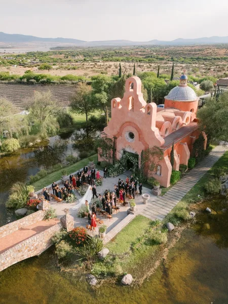 Outdoor wedding ceremony in a chapel courtyard with guests seated as the bride walks down the aisle in a long veil toward floral arch