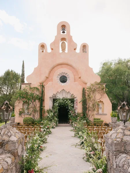 Ceremony aisle decor with a floral lined aisle, wooden chairs and stone lanterns leading to a stucco chapel entrance under blue sky
