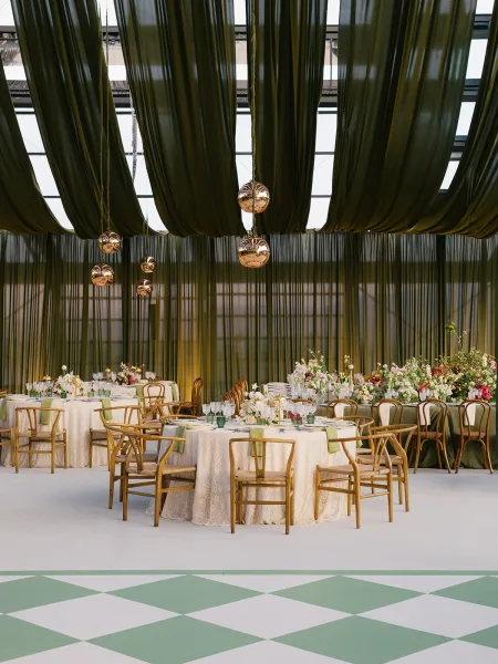 Reception tablescape with round wedding reception tables, lace linens, green napkins and tall florals under globe lights in a glass-ceiling conservatory