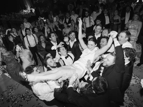 Wedding reception moment as guests lift the bride crowd surfing above the dance floor, confetti flying under reception lighting and greenery backdrop