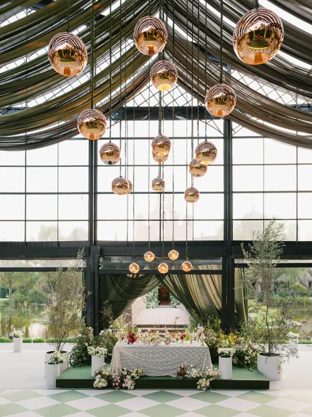 Ceremony altar in a greenhouse wedding ceremony with draped fabric ceiling, pendant lights, taper candles, and lush greenery-lined aisle runner