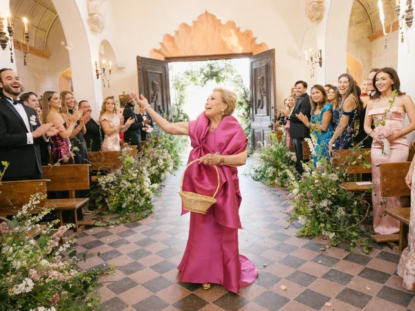 Flower girl entrance with a wicker basket, tossing petals down a greenery-lined chapel aisle as wedding guests cheer from wooden pews