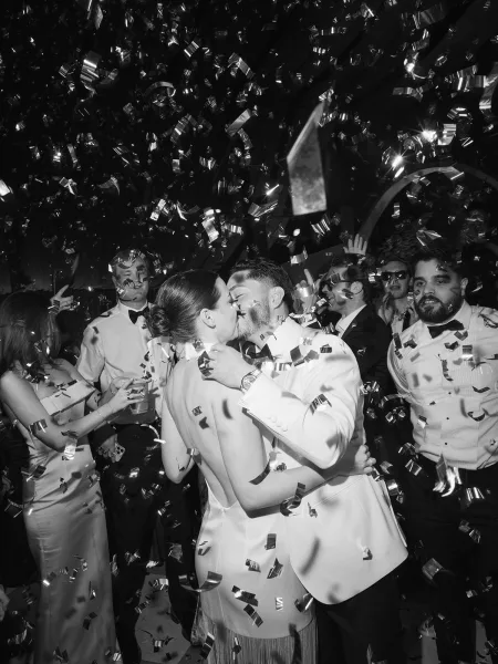 Wedding kiss as confetti rains down on the newlyweds, bride in a low-back gown and groom in a white tuxedo, guests filming in a dark reception space
