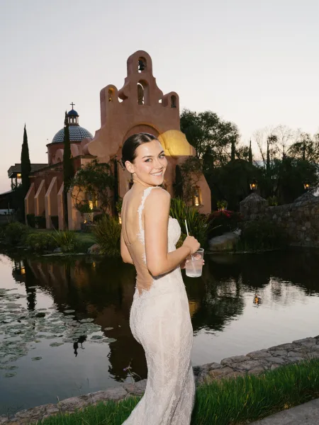 Bridal portrait of a bride looking back in a low back lace wedding dress, holding a drink by a twilight pond with water lilies near a church dome
