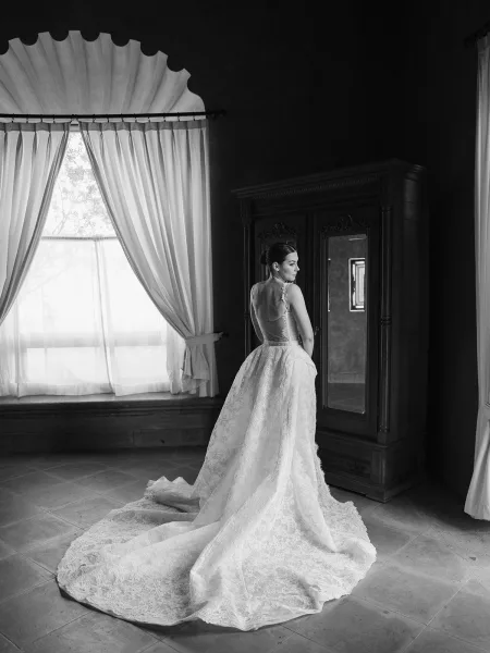 Bridal portrait in black and white of a bride looking over her shoulder, showing a low back lace gown by a window with sheer curtains
