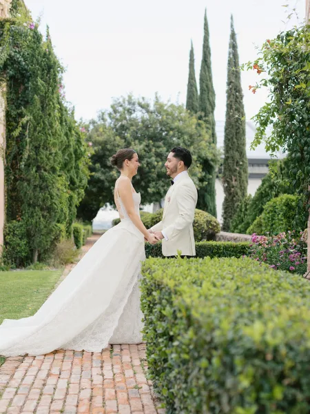Couple portrait of bride and groom holding hands, her lace gown train flowing on a brick garden path with tall cypress trees behind
