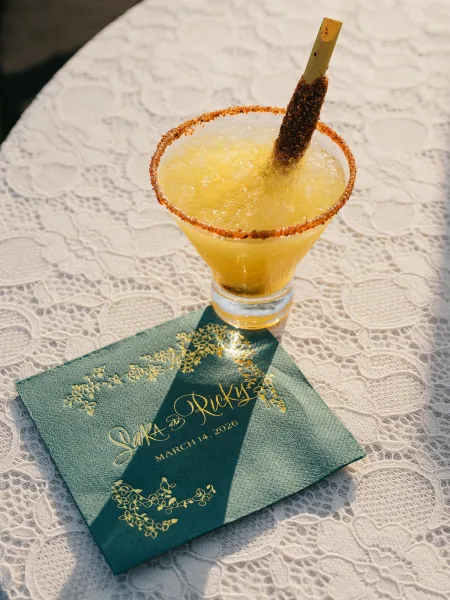 Wedding cocktail frozen margarita in a glass with chili salt rim and straw, on a sunlit lace tablecloth beside a gold foil napkin