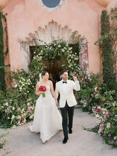 Wedding recessional with bride and groom walking out holding hands, bride with bouquet and veil under a pink floral arch by a stucco doorway