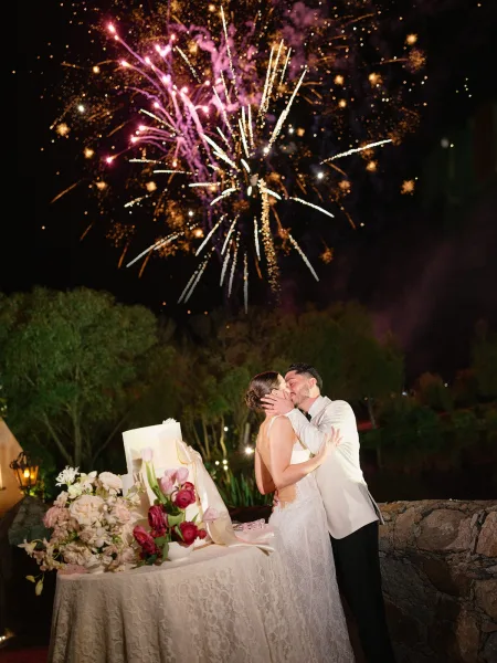 Wedding kiss as fireworks burst overhead, bride in lace dress and groom in a white suit beside a cake table, trees and stone wall behind