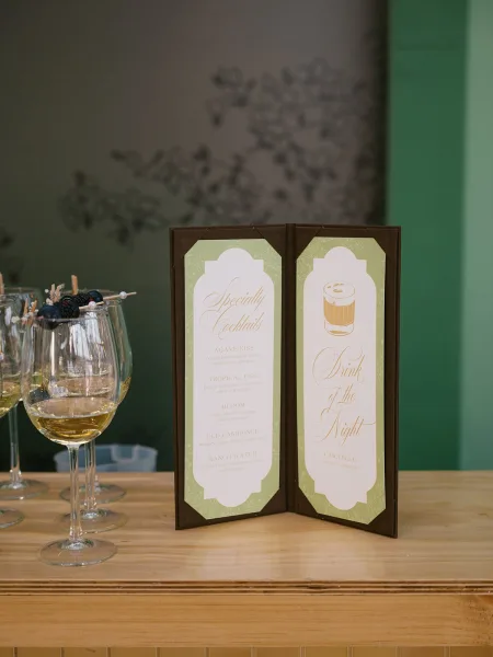 Wedding bar menu with signature cocktail menu in calligraphy on a folding card, styled with wine glasses and blackberries on a wood bar top