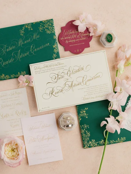 Invitation suite with emerald green wedding invitations and gold foil calligraphy, styled with blush flowers and rings on a linen backdrop
