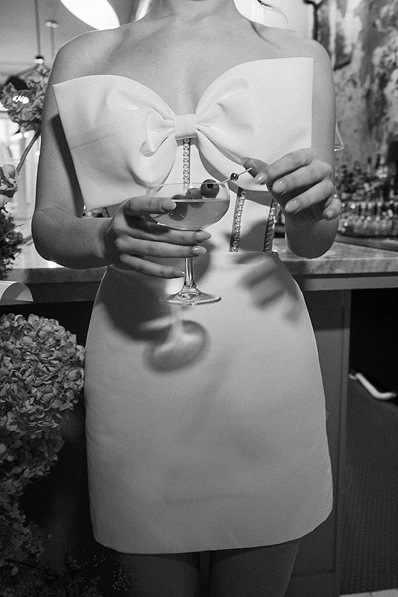 Wedding dress close-up of a short wedding dress with oversized bow bodice and pearl buttons as bride holds cherry-garnished coupe at bar counter
