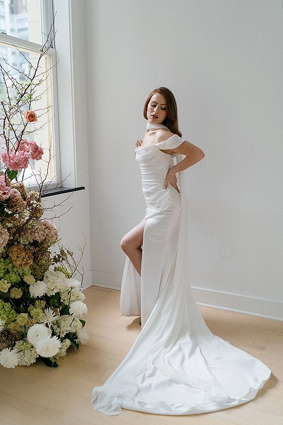 Bridal portrait in an off shoulder wedding dress with neck scarf and satin train, posed by a large window in natural light.