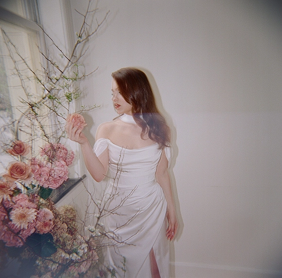Bridal portrait of a bride in an off shoulder wedding dress with neck scarf holding pink roses and carnations by a window on a white wall