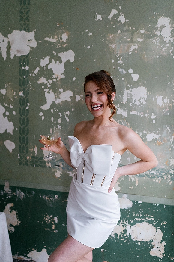 Bridal portrait of a smiling bride in a short wedding dress with an oversized bow, holding a champagne coupe against a distressed wall backdrop