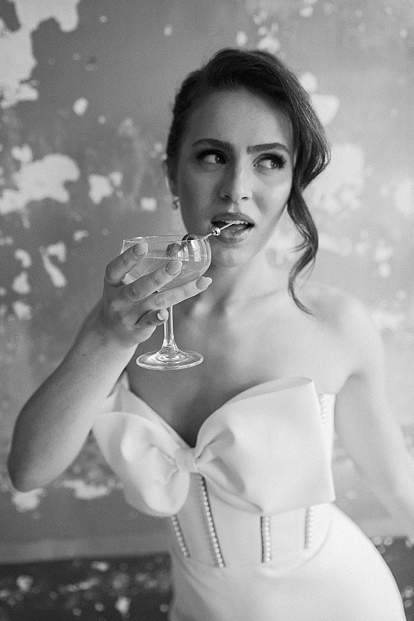 Bridal portrait of a bride holding cocktail in a strapless bow wedding dress with pearl corset bodice, posed by a peeling textured wall