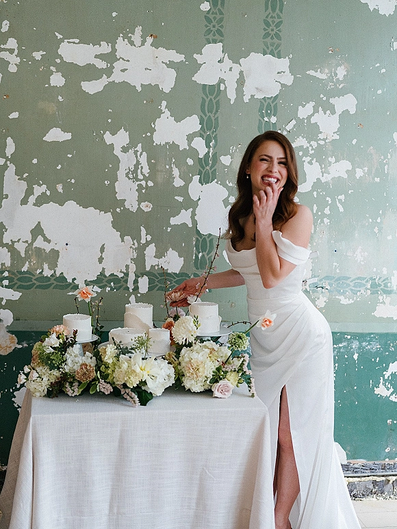 Bridal portrait of a bride with wedding cake at a white linen cake table, wearing an off-shoulder slit gown before a distressed wall backdrop