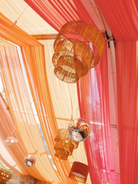 Wedding ceiling decor with colorful fabric ceiling draping, rattan pendant lampshades, disco balls, and string lights under a daylight tent canopy