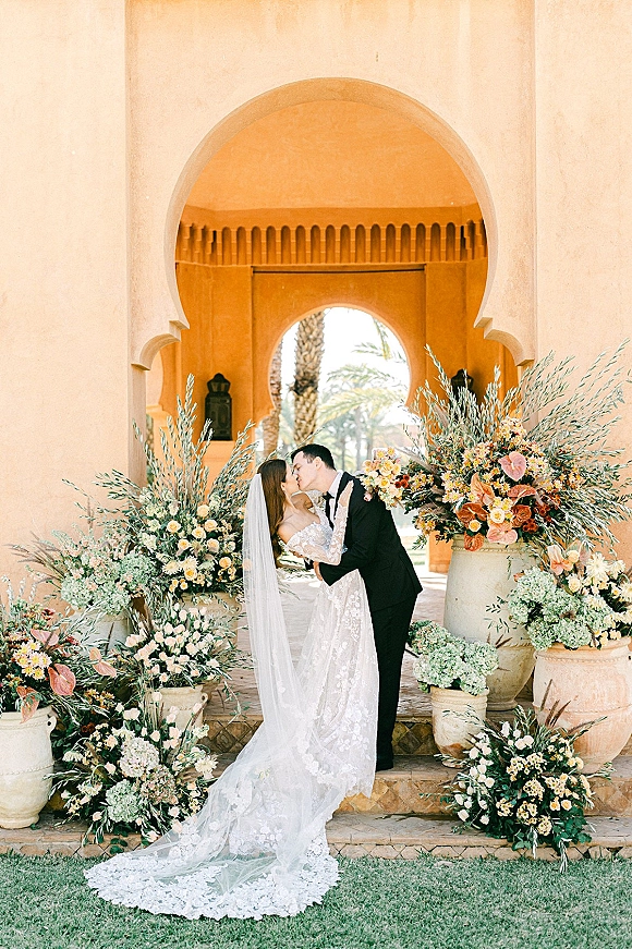 Wedding kiss portrait of bride and groom kissing under an arched doorway, lace dress and long veil, flanked by white florals in urns