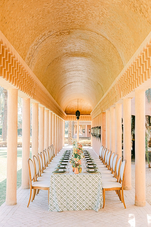 Reception tablescape on a long banquet table setup with patterned tablecloth, green plates, florals and taper candles under an arched colonnade courtyard