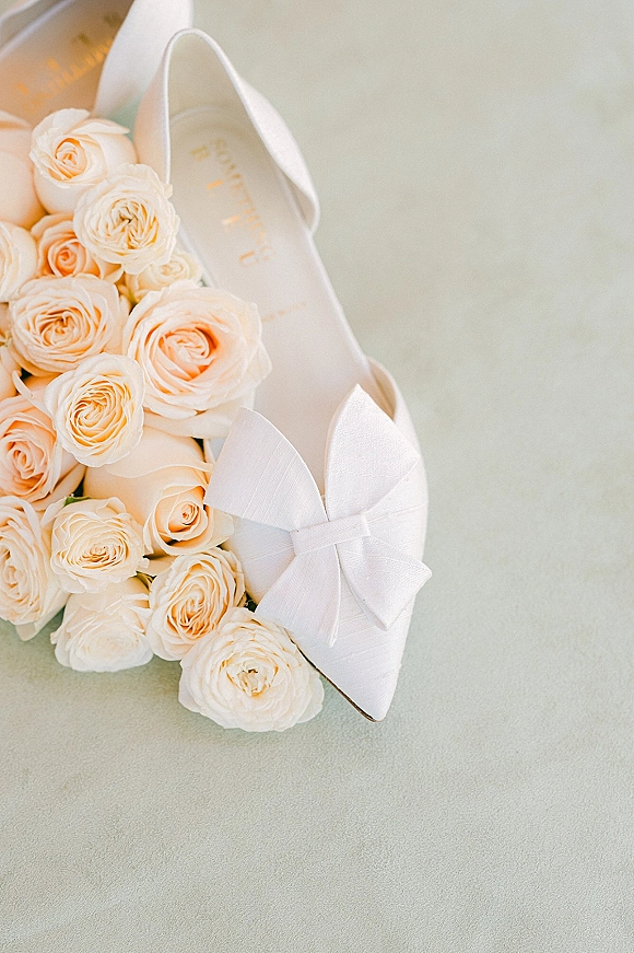 Bridal shoes in a wedding shoes flat lay, white pointed-toe heels with bow detail beside ivory roses on a soft gray surface