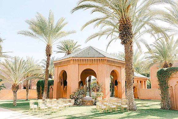 Outdoor ceremony setup with garden ceremony chairs in rows, aisle steps lined with floral urns and arrangements under a stucco arch and palm trees.