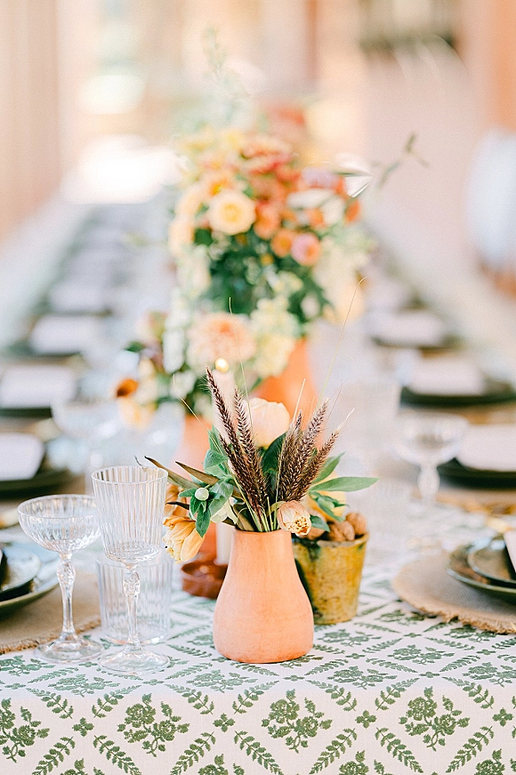 Reception tablescape with terracotta vase centerpieces, greenery and wheat stems, taper candles, and patterned linens on a long banquet table indoors