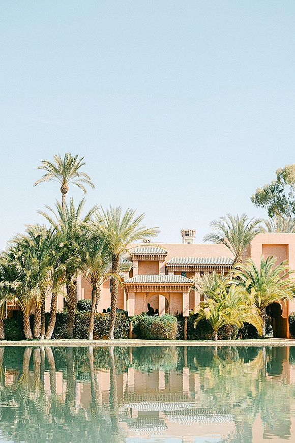 Wedding venue exterior with a tiled roof and arches, palm trees, and a clear water reflection by a pond under blue sky