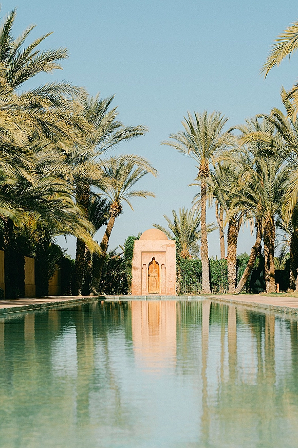 Wedding venue pool with a reflecting pool leading to a domed pavilion and arched doorway, framed by palm trees and hedges under blue sky