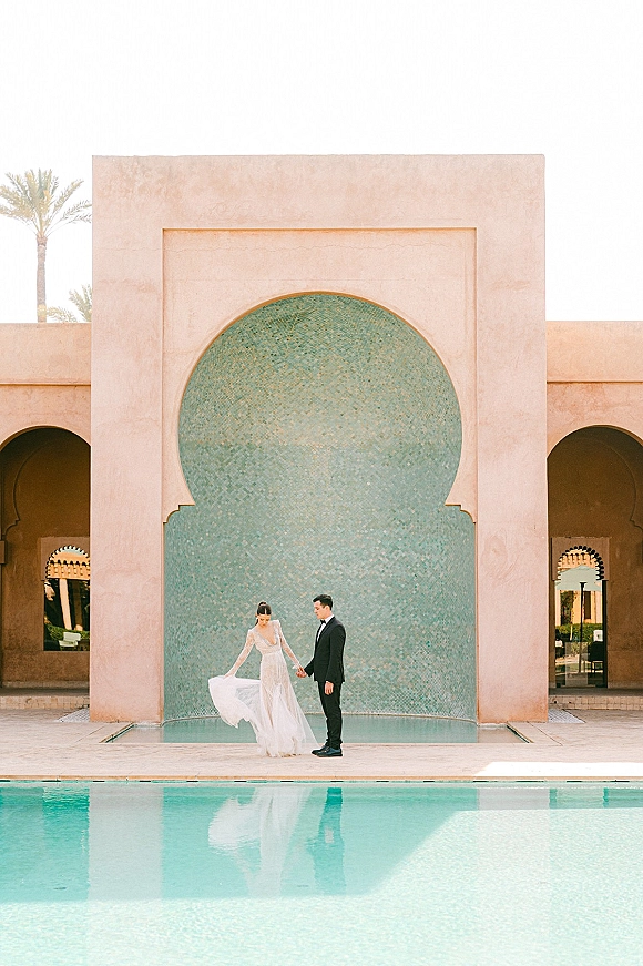 Couple portrait of bride and groom holding hands by a reflecting pool, her veil trailing near an arched mosaic tile courtyard facade