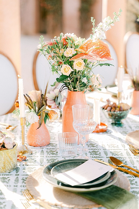 Reception tablescape with wedding table centerpiece in a peach ceramic vase, white taper candles in brass holders, patterned linens, and fruit bowl indoors
