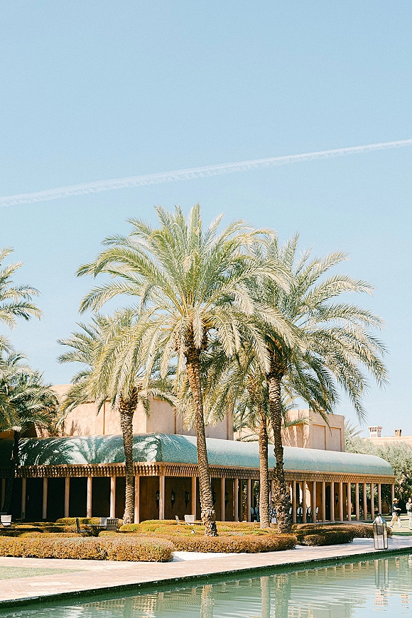 Wedding venue exterior with palm trees framing a columned entrance, hedges and lanterns lining the walkway beside a reflecting pool under blue sky