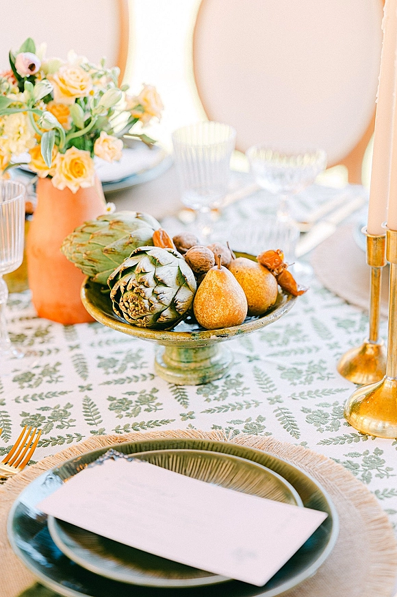 Reception tablescape with a wedding place setting on a patterned tablecloth, brass taper candles, green plates, and fruit centerpiece on tabletop
