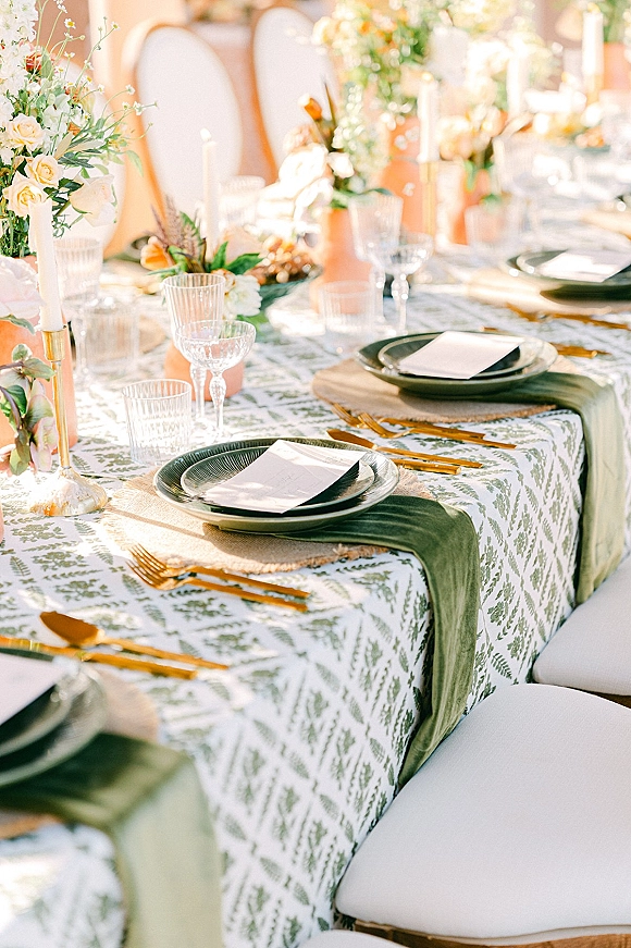 Reception tablescape with wedding table setting on a botanical print tablecloth, sage runner, floral centerpiece, and taper candles in outdoor light