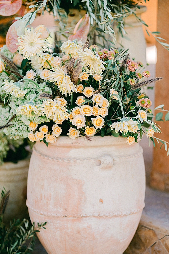 Wedding floral arrangement in a stone urn with yellow roses, cream dahlias, succulents and eucalyptus on a stone floor by a wooden structure