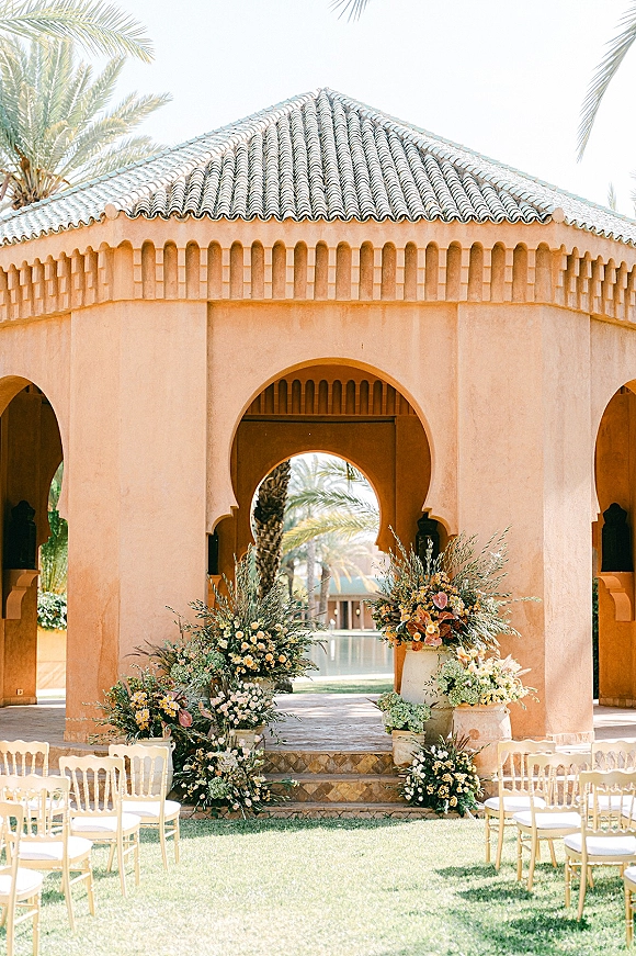 Ceremony setup for an outdoor wedding ceremony with white chairs, floral urns and greenery on stone steps beneath an arched pavilion, palm trees beyond