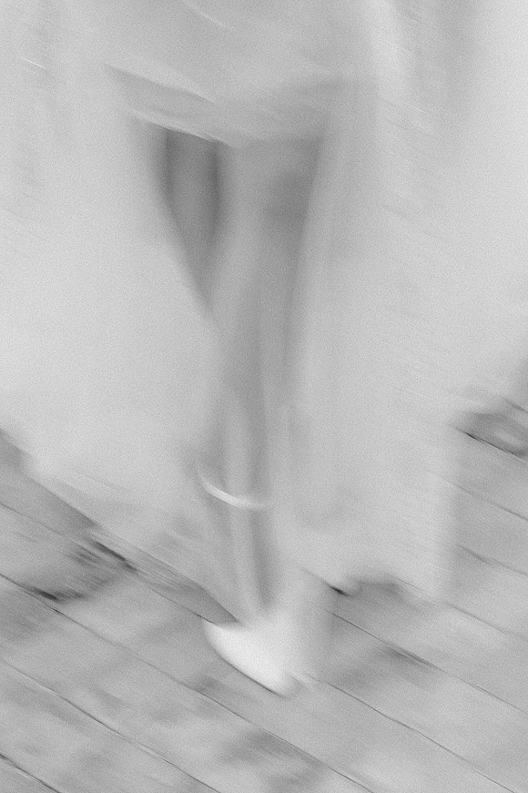 Wedding dress detail with bridal shoes as white pointed-toe heels peek from the gown hem in a black-and-white motion blur on wood floor
