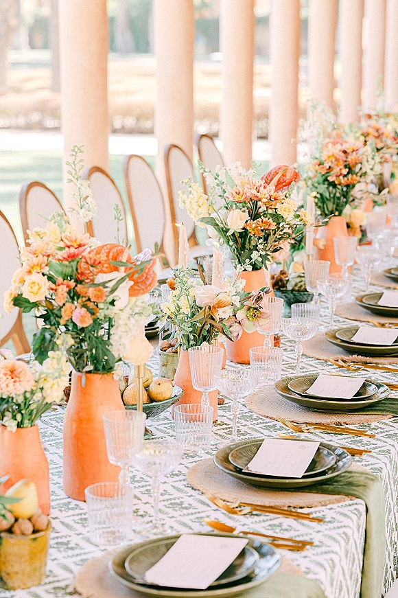 Reception tablescape with long reception table decor, orange floral centerpieces in peach ceramic vases, taper candles, patterned linens on a patio