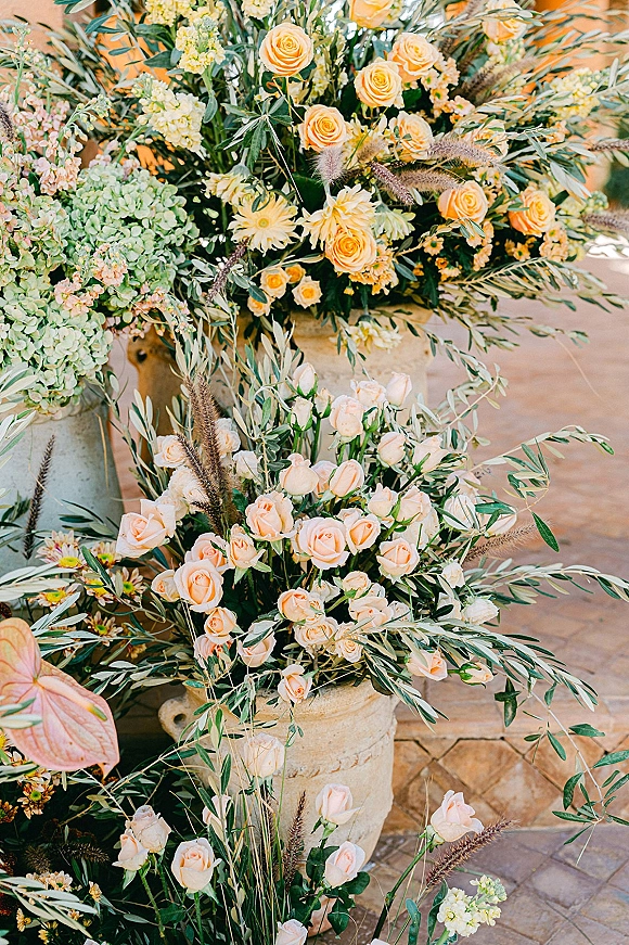 Wedding floral arrangement of peach and yellow roses with hydrangea on a stone pedestal, framed by garden patio greenery and grasses