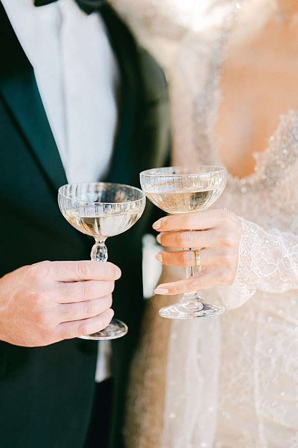 Wedding toast as bride and groom clink champagne coupes, rings visible on hands with lace sleeve and tuxedo in soft blur background