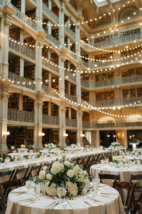 Reception tablescape with round wedding table setup, ivory linens, white florals and greenery, taper candles under bistro string lights in a grand atrium