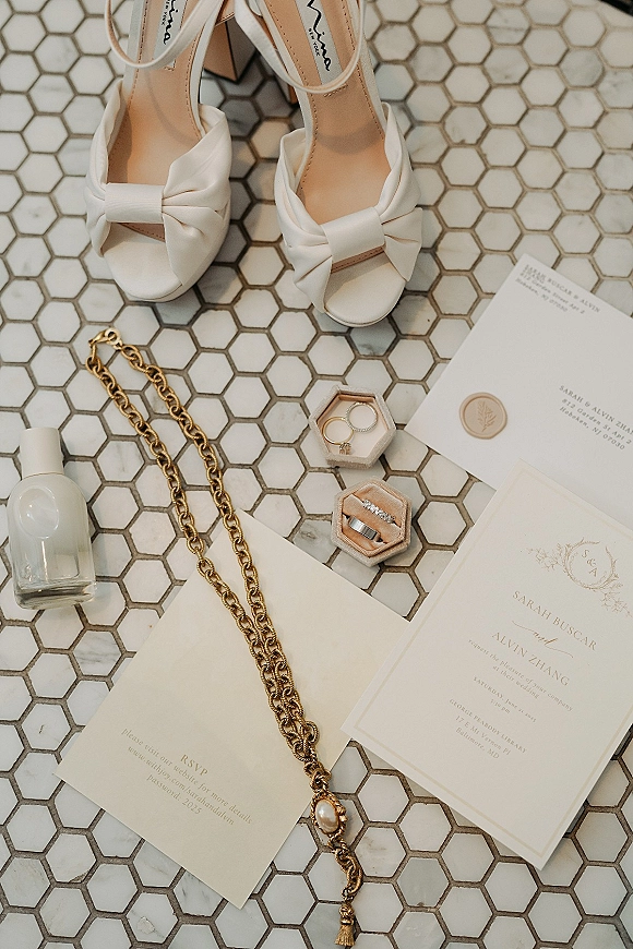 Wedding flat lay with wedding invitation suite, RSVP and envelopes, wax seal, rings in boxes, pearl pendant, perfume, and bow heels on white hexagon tile floor